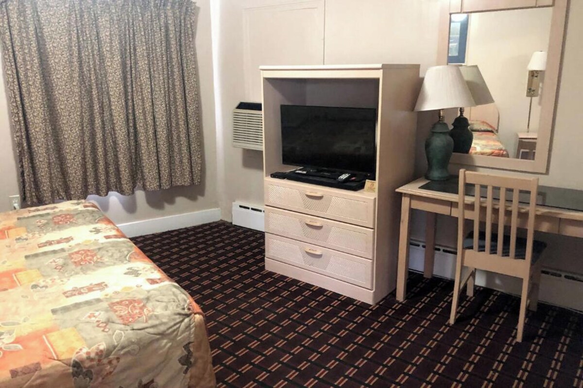 A hotel room with a bed, a desk with a lamp, a television, and a dresser.