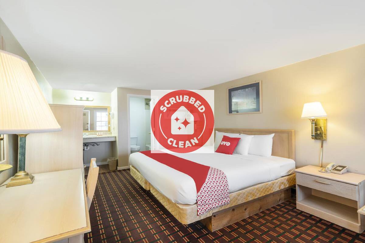 A hotel room with a bed, bedside table, lamp, and a red sign indicating the room is scrubbed clean.