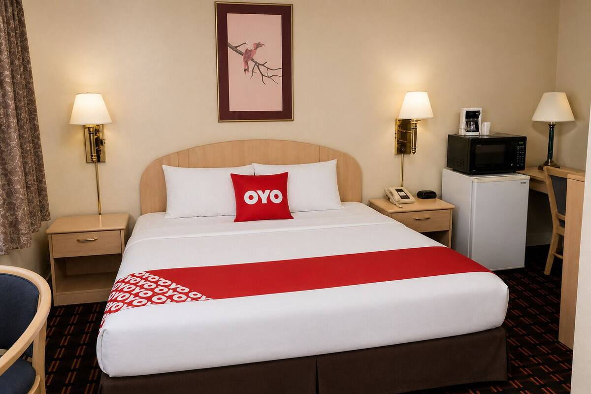 A hotel room with a bed, two bedside tables, a desk with a chair, a microwave, a refrigerator, and a framed picture on the wall.