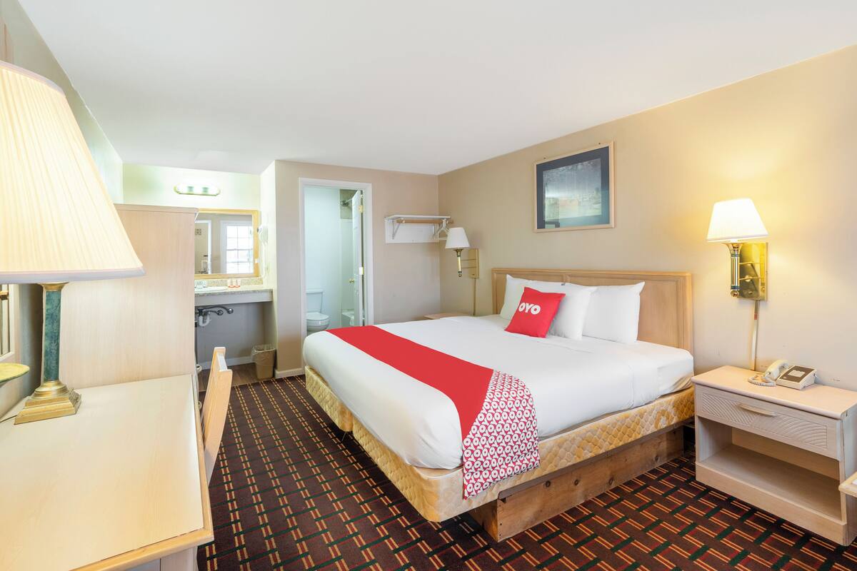 A hotel room with a bed, bedside table, lamp, and a view of the bathroom.