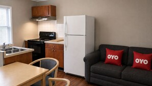 A compact kitchen with a stove, sink, and refrigerator, a dining table with chairs, and a gray sofa with red pillows. - OYO Hotel Starlite Seneca Falls (Seneca Falls)