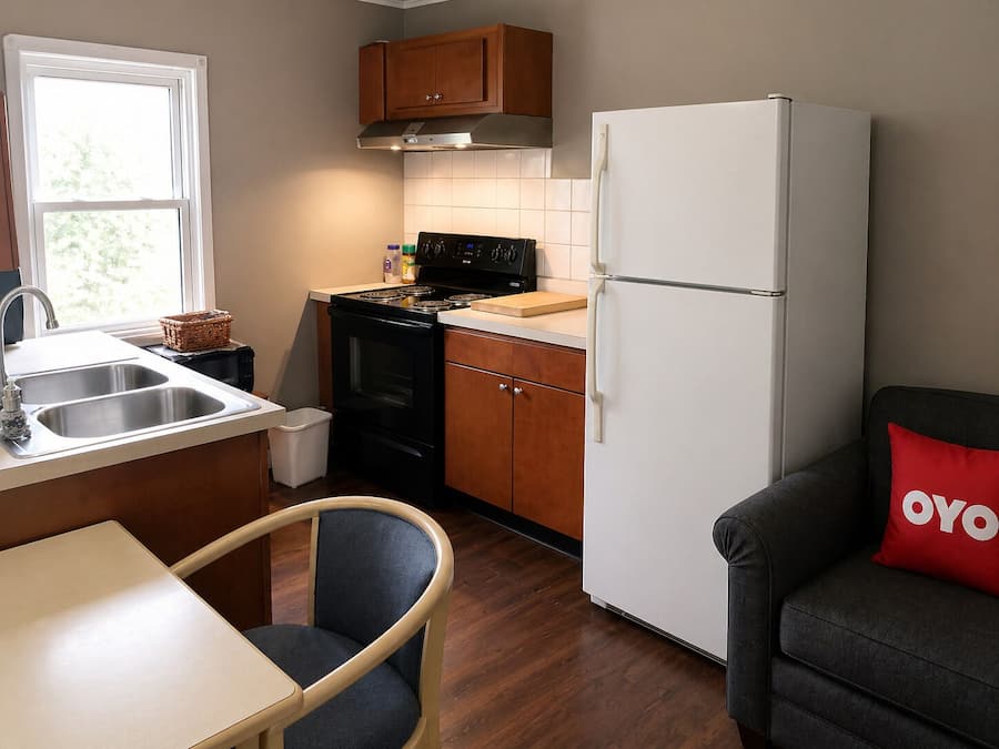 Cocina compacta con estufa, pileta y refrigerador, alacenas de madera, una ventana y un comedor con una silla.