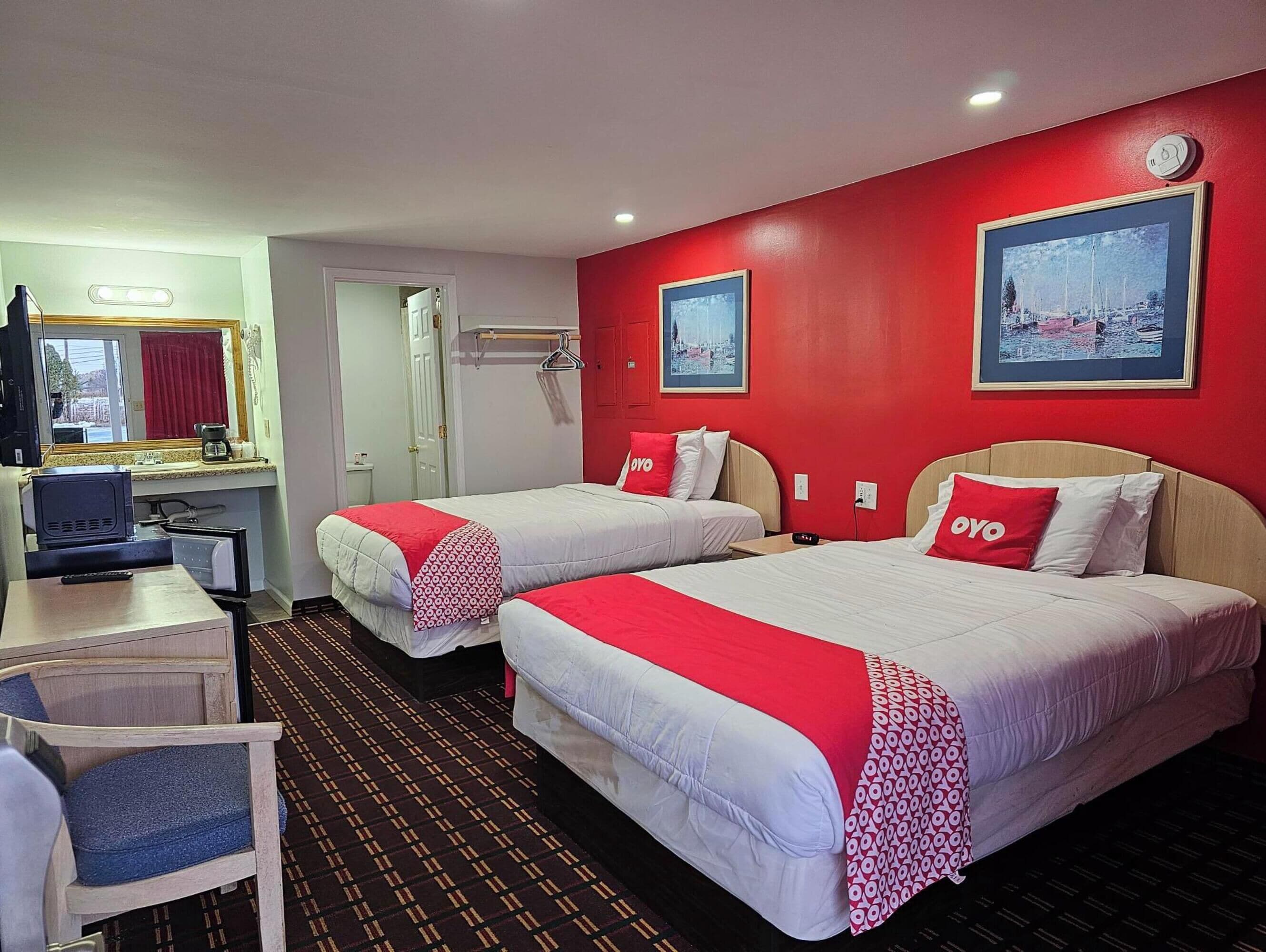 Ein Hotelzimmer mit zwei Betten, einem Schreibtisch, einem Stuhl und einer roten Wand mit zwei gerahmten Bildern.