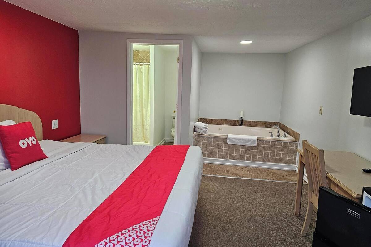 A hotel room with a bed, a desk, a chair, a bathroom with a bathtub, and a television on the wall.