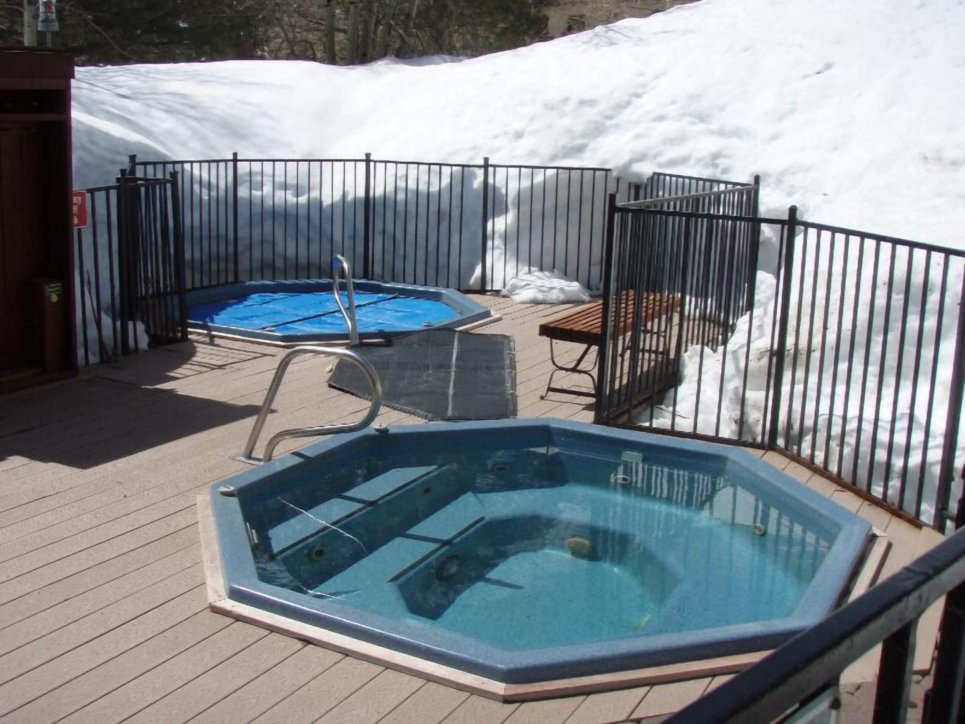 Un bain à remous sur une terrasse en bois, entouré d’une clôture métallique, le tout noyé dans la neige.