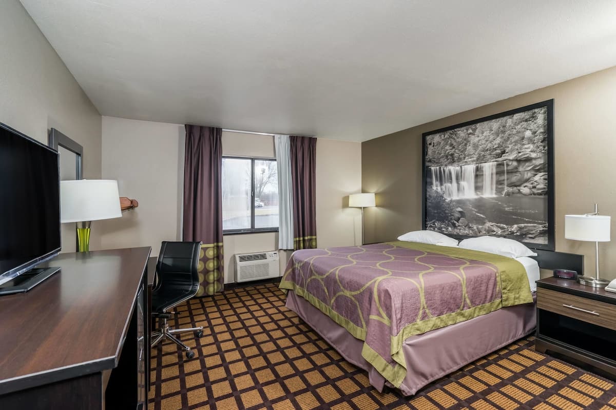 A hotel room with a bed, a desk with a TV, a chair, a window with curtains, a wall-mounted TV, and a painting of a waterfall.