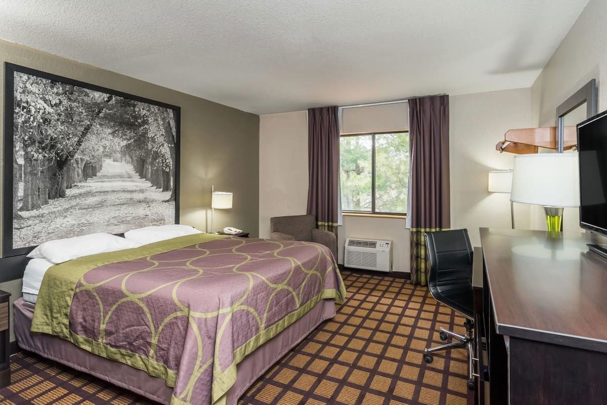 A hotel room with a bed, a desk with a television, a chair, a window with curtains, and a large framed picture on the wall.
