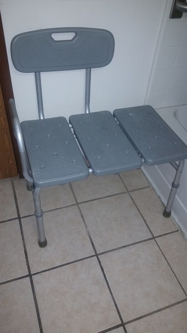 Un banco de metal gris con respaldo y cuatro patas, sobre un piso de baldosas.