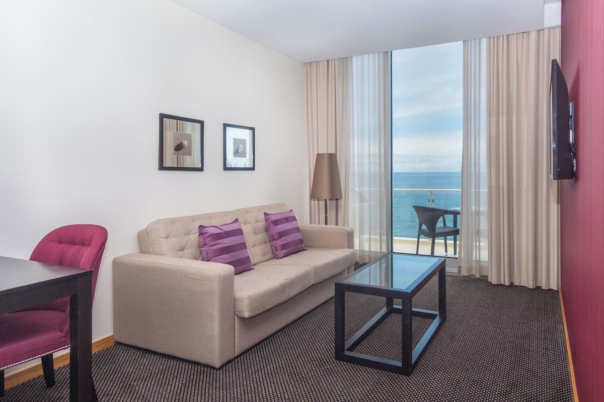 Ein Hotelzimmer mit einer Couch, einem Glastisch, einem roten Sessel und Meerblick durch große Fenster.