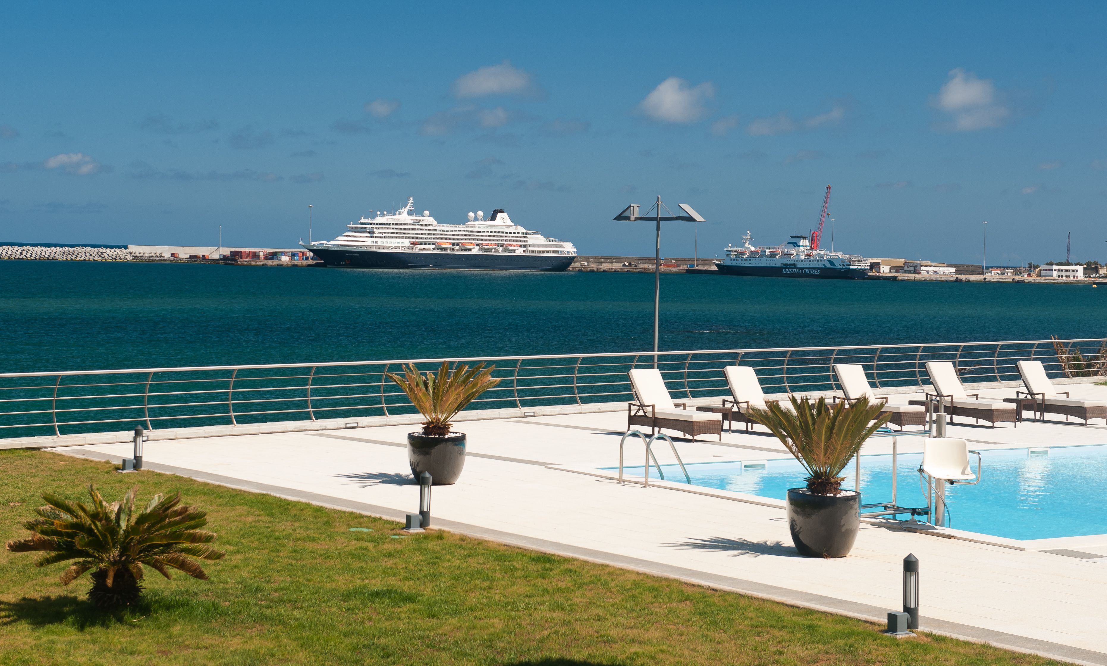 A pool area with lounge chairs and potted plants, overlooking a body of water with cruise ships.