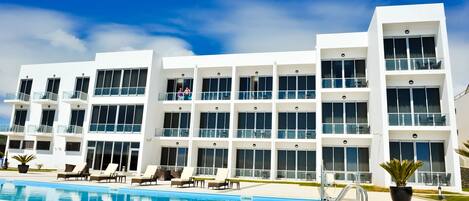 A modern hotel with multiple balconies, a swimming pool, and lounge chairs.
