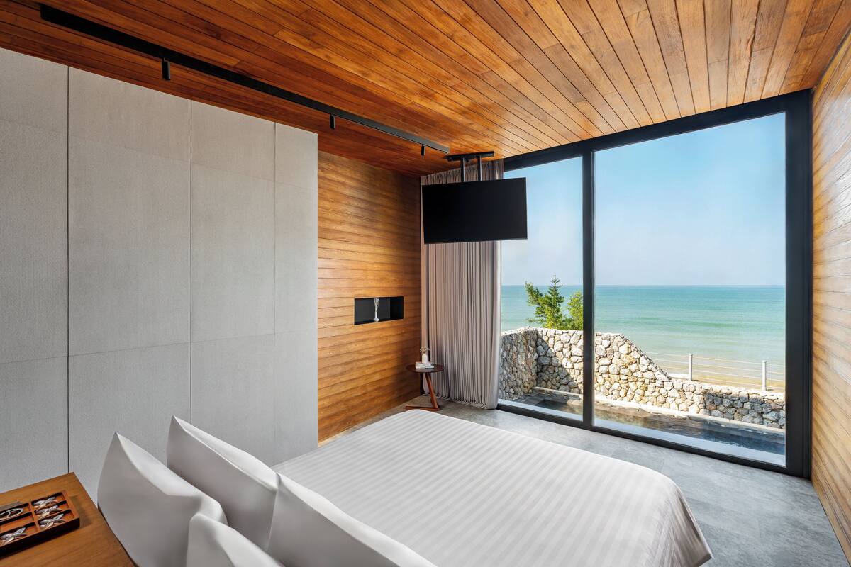 A modern bedroom with a large bed, a TV mounted on the wall, and a view of the sea through floor-to-ceiling windows.