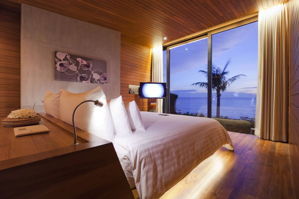 A bedroom with a bed, a bedside table, a TV, and a view of the ocean.