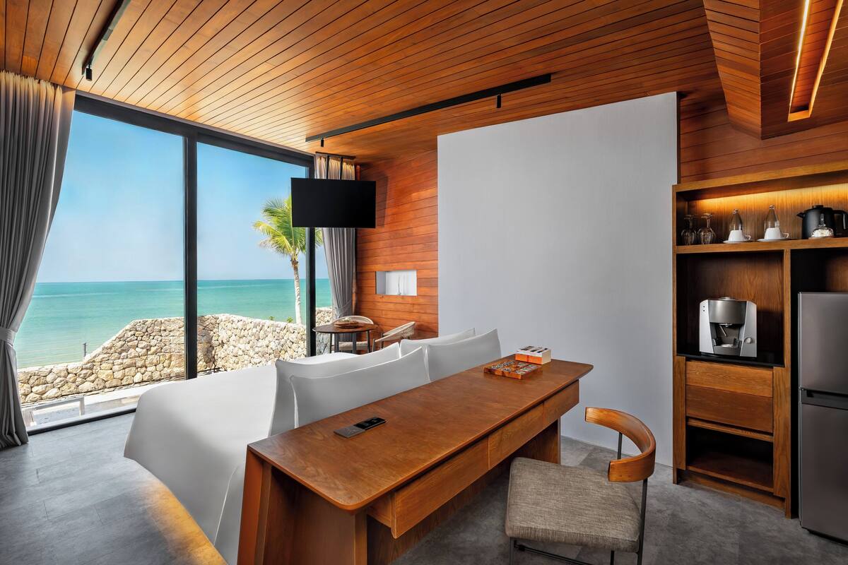A modern room with a wooden ceiling, a large window overlooking the sea, a dining table with chairs, a flat-screen TV, and a kitchen area with a coffee machine.