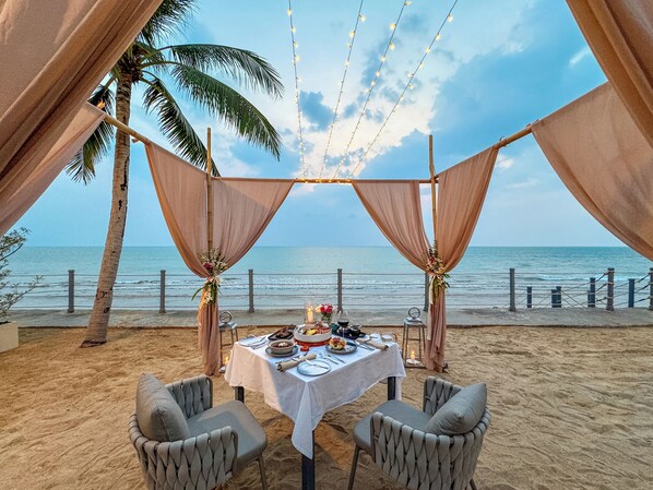 A beachside dining setup with a table and chairs, draped with fabric, overlooking the ocean.