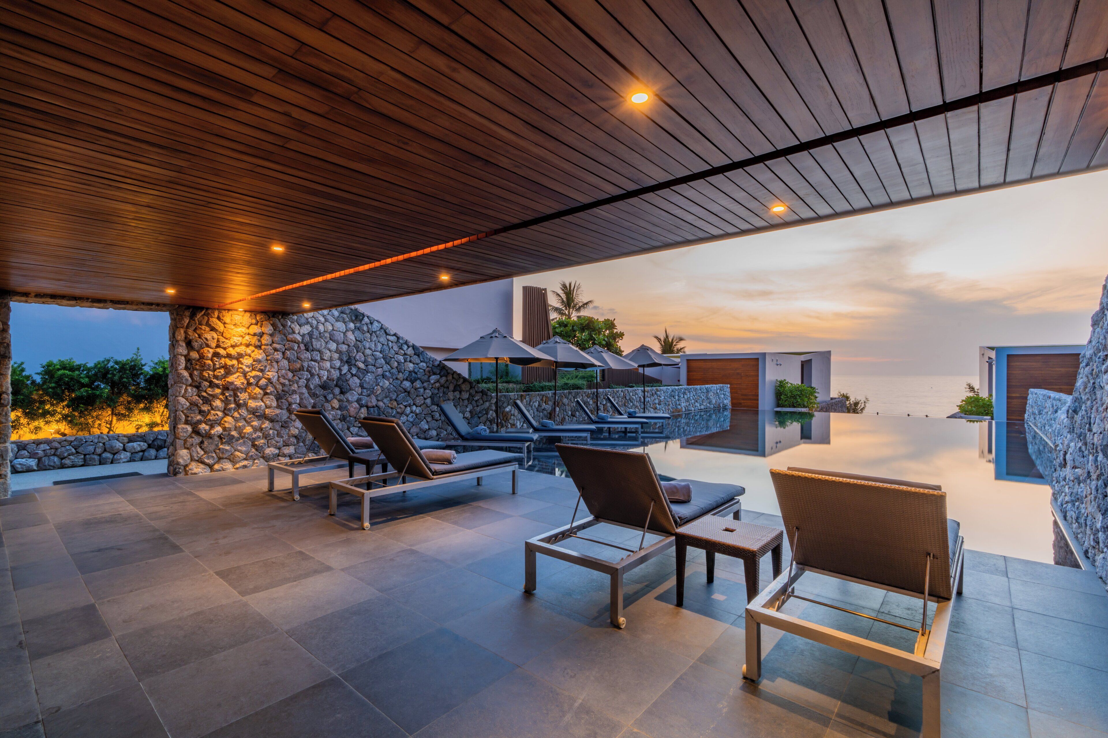 A covered outdoor area with lounge chairs, a stone wall, and a view of the ocean at sunset.