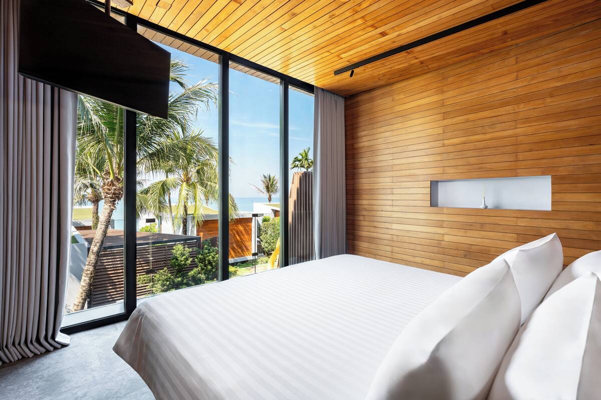 A modern hotel room with a large bed, wooden panel walls, and a view of palm trees and a pool.