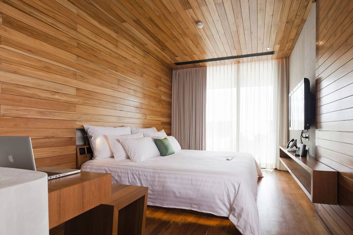 A modern bedroom with a wooden ceiling, a bed with white bedding, a desk with a laptop, and a TV mounted on the wall.