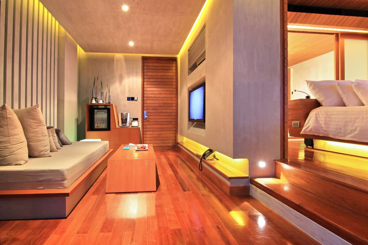 A modern hotel room with a wooden floor, a sofa, a TV, and a bed with a wooden headboard.