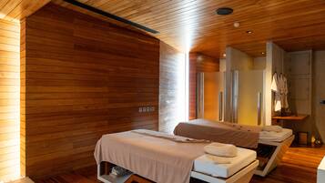 A modern spa room with wooden walls and ceiling, two beds with towels, a mirror, and a small table with towels.