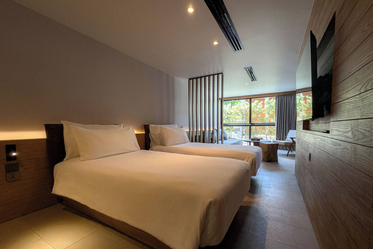 A modern hotel room with two beds, a large window, a TV mounted on the wall, and a small table.