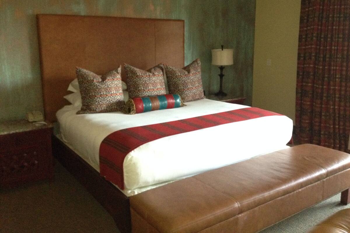 Une chambre d’hôtel avec un grand lit, une tête de lit en bois, des coussins à motifs, une lampe de chevet et un banc en cuir marron.