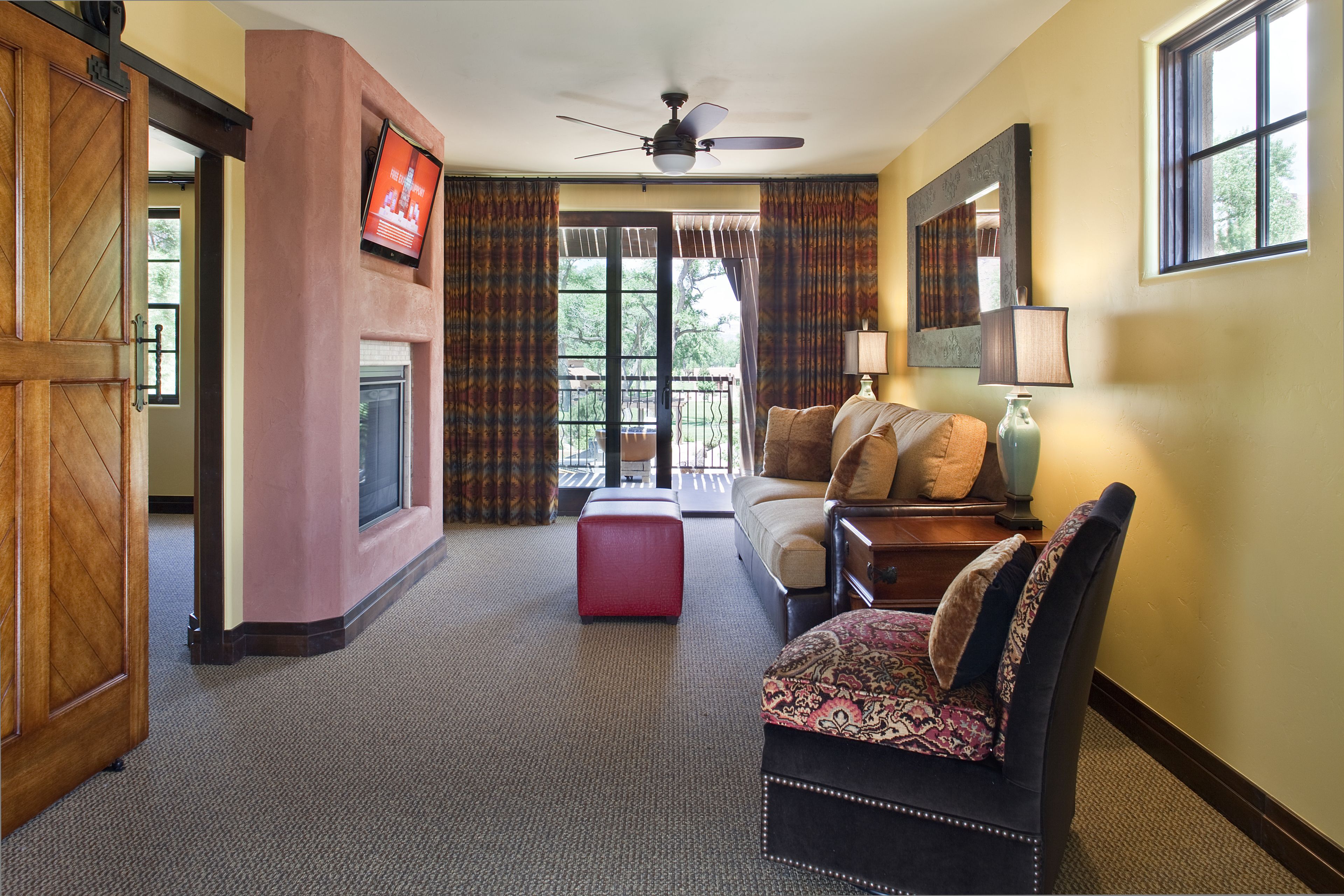 Une chambre d’hôtel avec une cheminée, un canapé, un pouf rouge, un ventilateur de plafond et une vue sur l’extérieur.