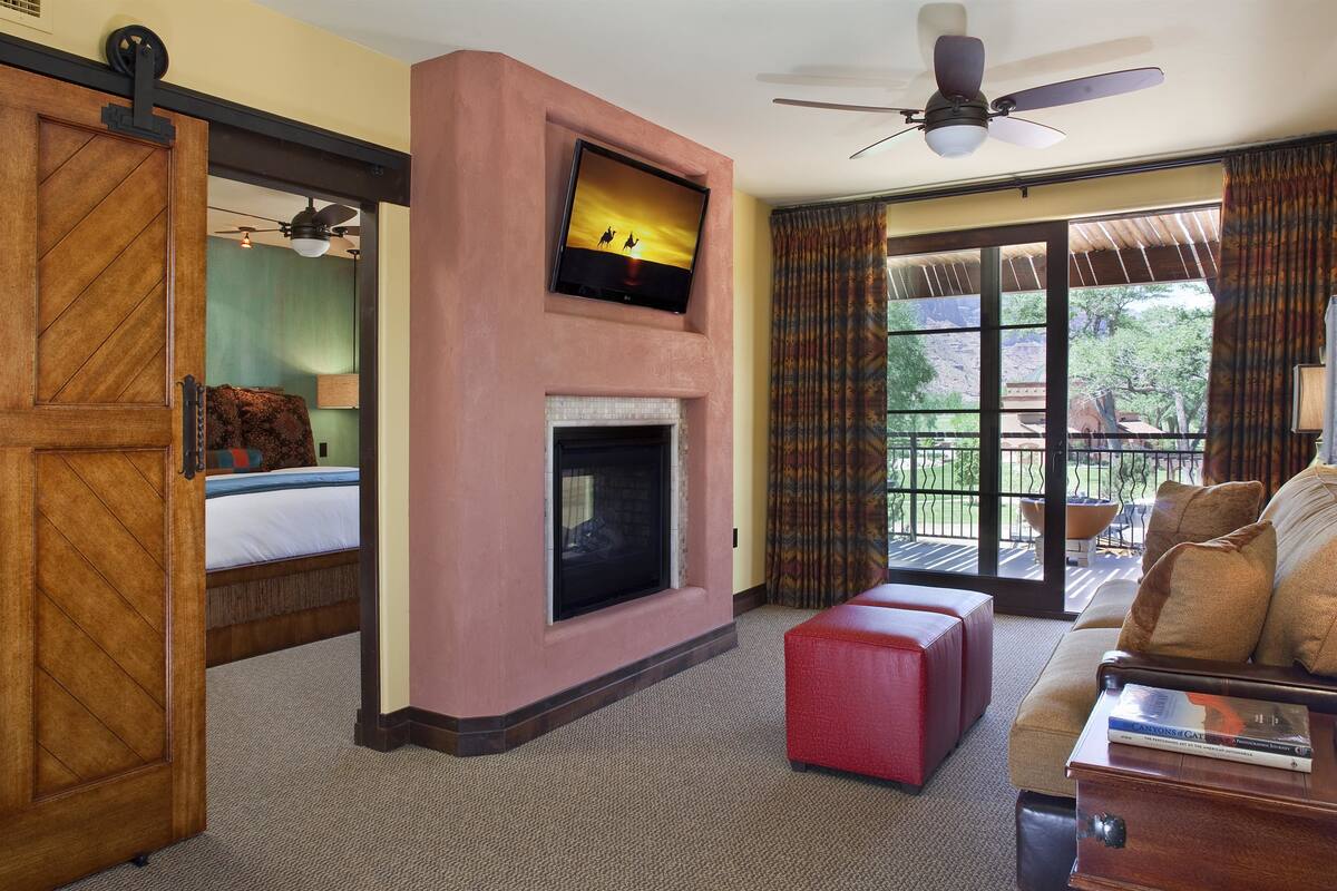 Une chambre d’hôtel avec une cheminée, un téléviseur fixé au-dessus, un balcon donnant sur l’extérieur et un pouf rouge.
