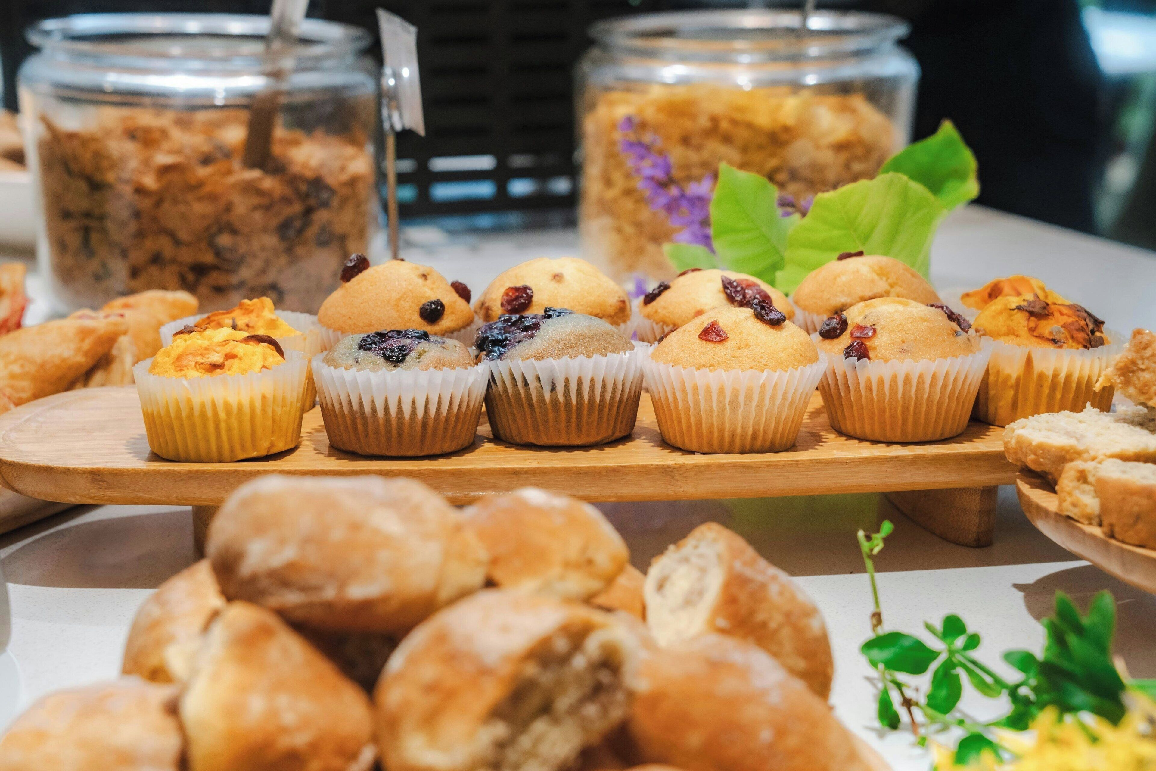 A variety of baked goods on a wooden board, including muffins and pastries, with two jars of granola in the background.
