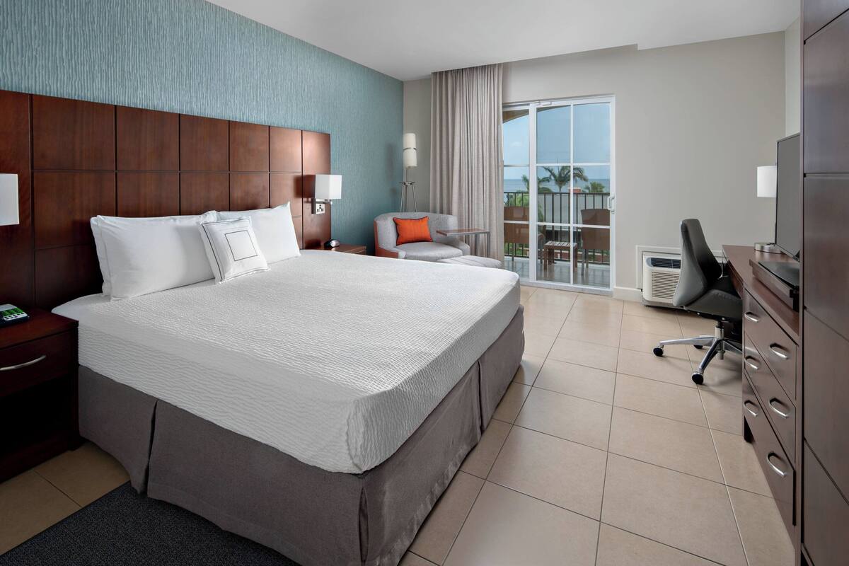 A hotel room with a large bed, a desk, a chair, a TV, and a balcony with a view.