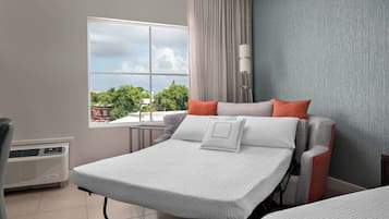Ett hotellrum med en säng, en stol, ett litet bord och ett fönster med utsikt över träd och byggnader.