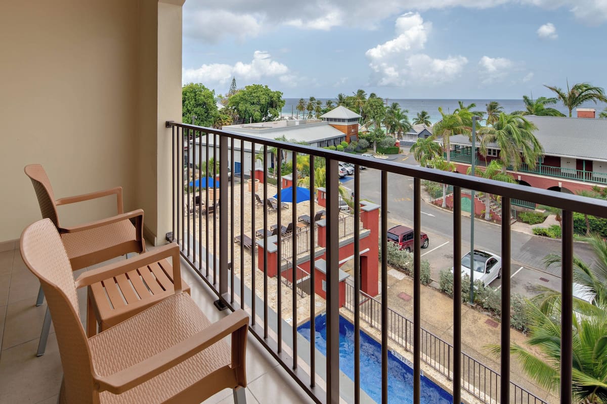 A balcony with two wicker chairs, a view of a swimming pool, and a street with parked cars and buildings.