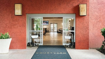A hotel entrance with a red textured wall, a 'COURTYARD' doormat, and a reception area visible through the glass doors.