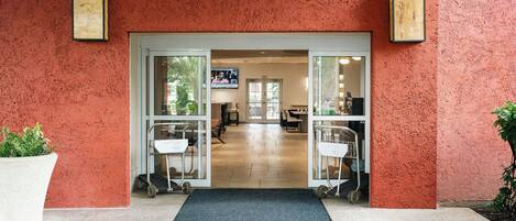 Entrada de un hotel con un muro rojo texturizado, un felpudo con la leyenda 'COURTYARD' y un área de recepción visible a través de las puertas de cristal.