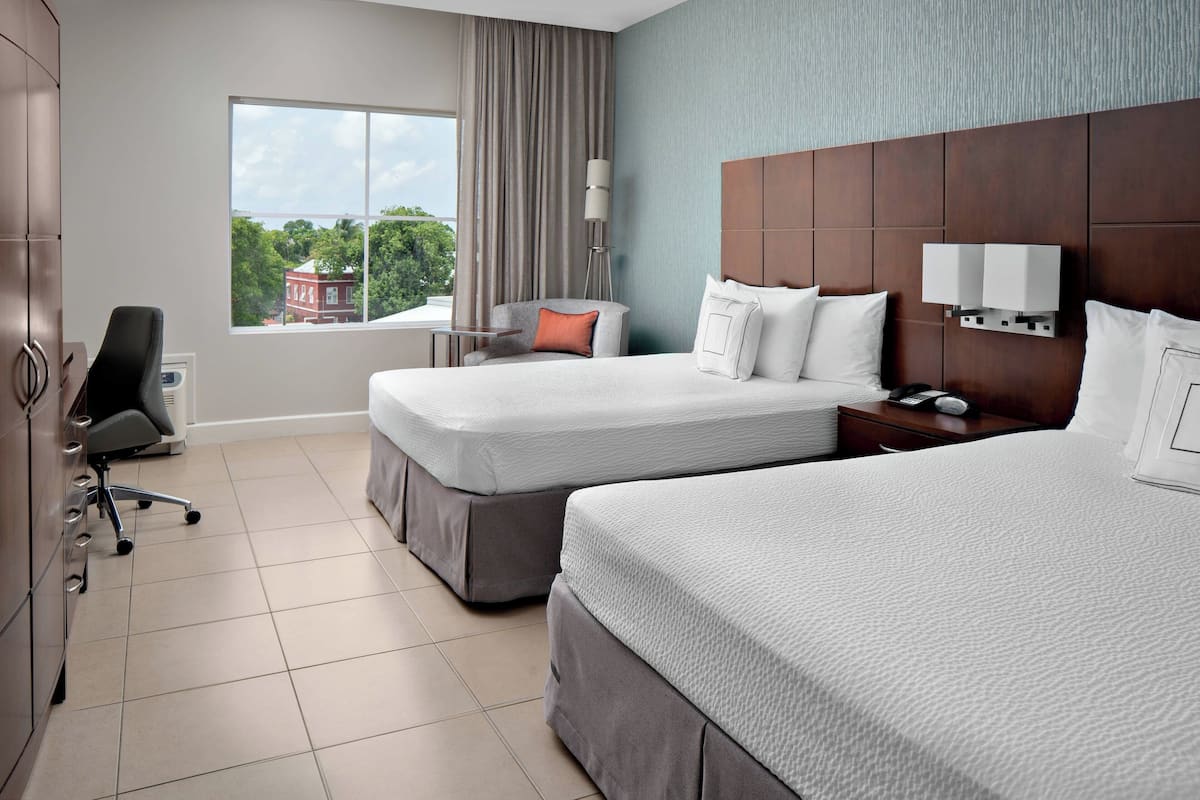 A hotel room with two beds, a desk, a chair, and a window with a view of trees and a building.