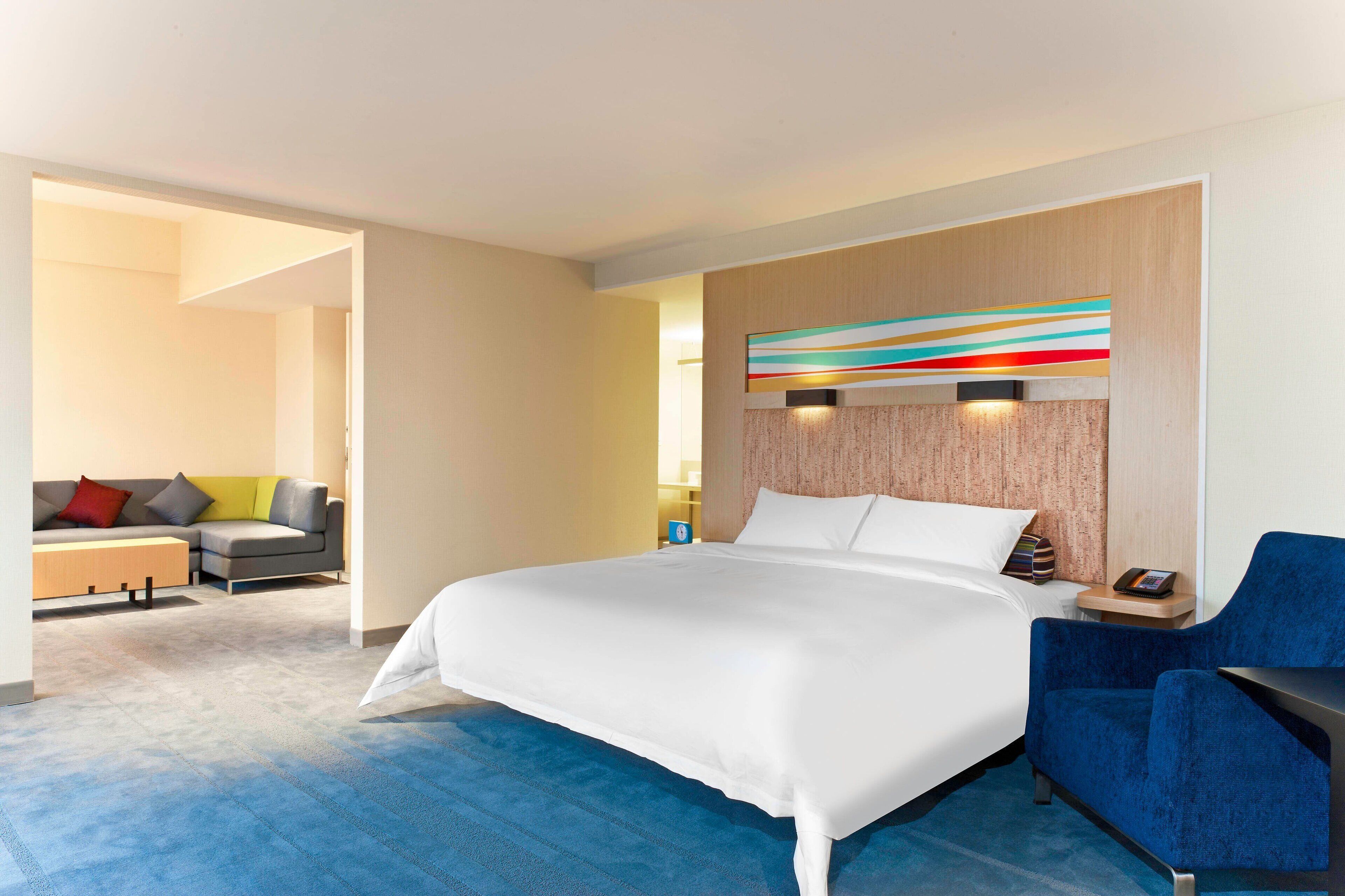 Una habitación de hotel moderna con una cama grande, un sillón azul y un conjunto de sofás al fondo.
