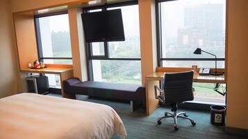 A hotel room with a bed, desk, chair, TV, and large windows offering a city view.