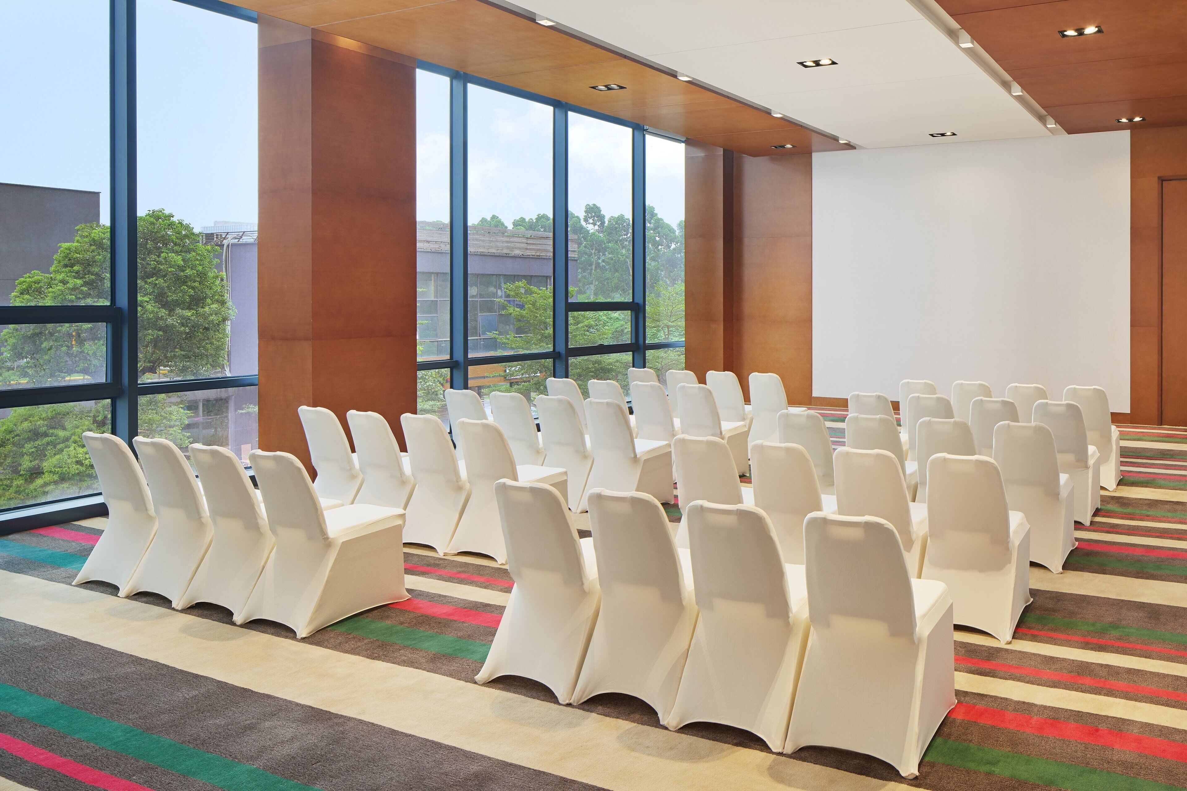 Un salón de conferencias con filas de sillas blancas, ventanas amplias y suelo alfombrado.