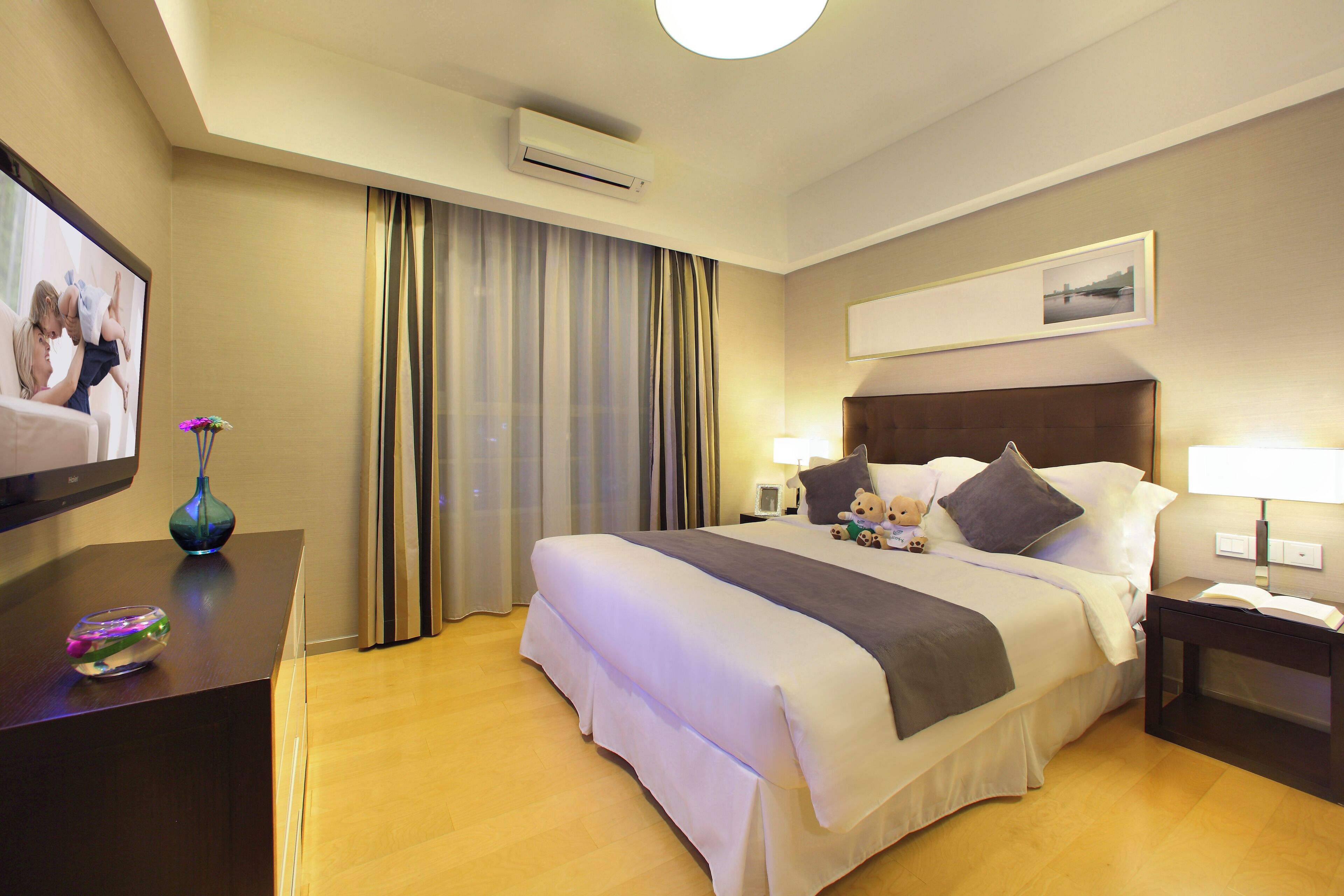 A hotel room with a large bed, a flat-screen TV, a bedside table with a lamp, and a window with curtains.