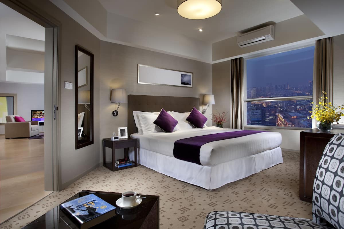 Ein Hotelzimmer mit einem großen Bett, einem Schreibtisch mit einer Kaffeetasse, einem Sessel und Blick auf die beleuchtete Stadt bei Nacht.