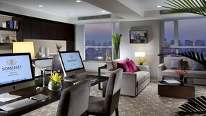 Una habitación de hotel moderna con escritorio, dos monitores, un sofá y un ventanal con vista a la ciudad.