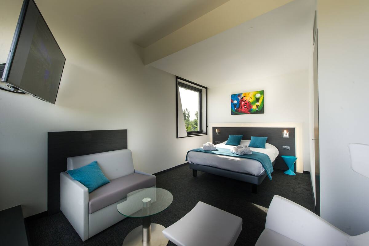 A hotel room with a bed, a sofa, a glass table, and a TV mounted on the wall.