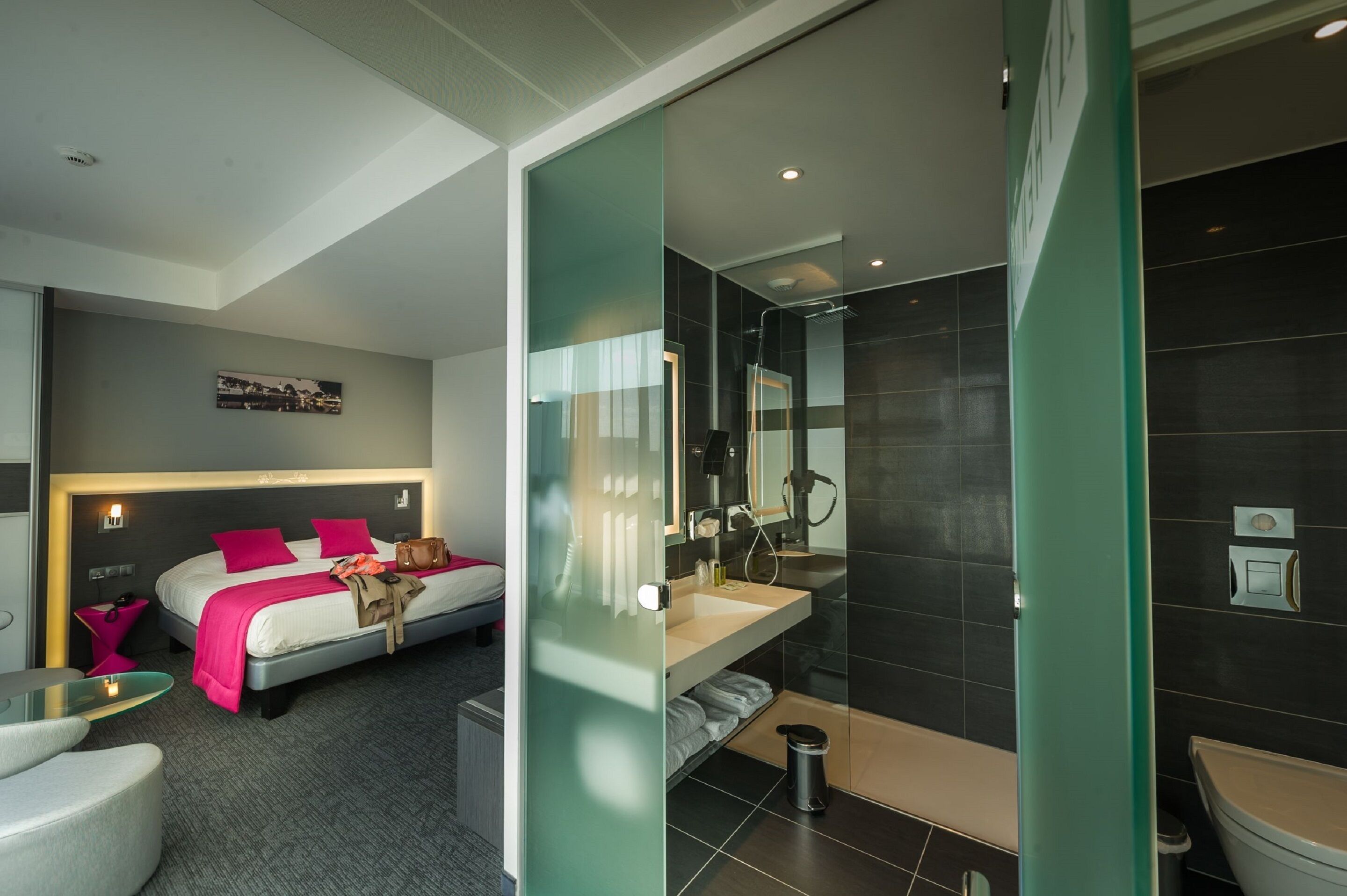 A modern hotel room with a bed, bedside table, and a glass partition separating the bedroom from the bathroom.