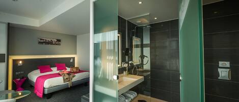 A modern hotel room with a bed, bedside table, and a glass partition separating the bedroom from the bathroom.