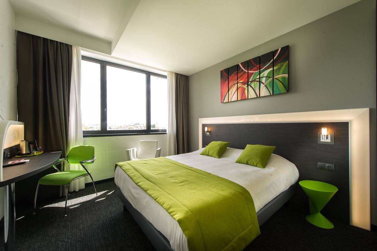 A hotel room with a large bed, a desk with a chair, a window with curtains, and a painting on the wall.