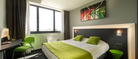 A hotel room with a large bed, a desk with a chair, a window with curtains, and a painting on the wall.