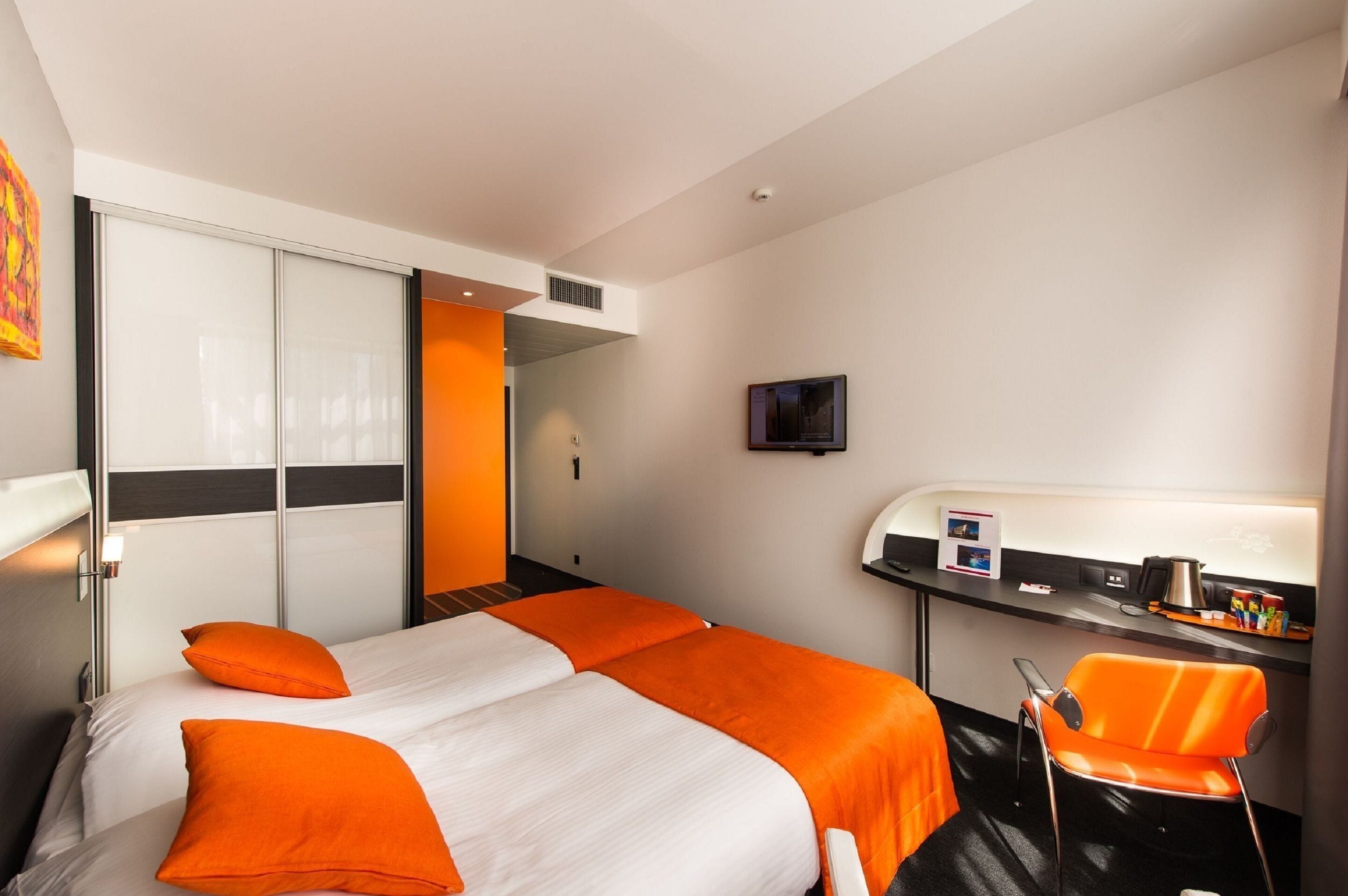 A hotel room with a bed, an orange chair, a desk with a computer, and a TV.