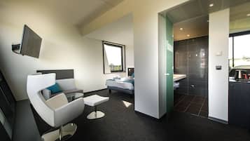 A modern hotel room with a sofa, a small table, a bed, a bathroom with a sink, and a large window providing natural light.