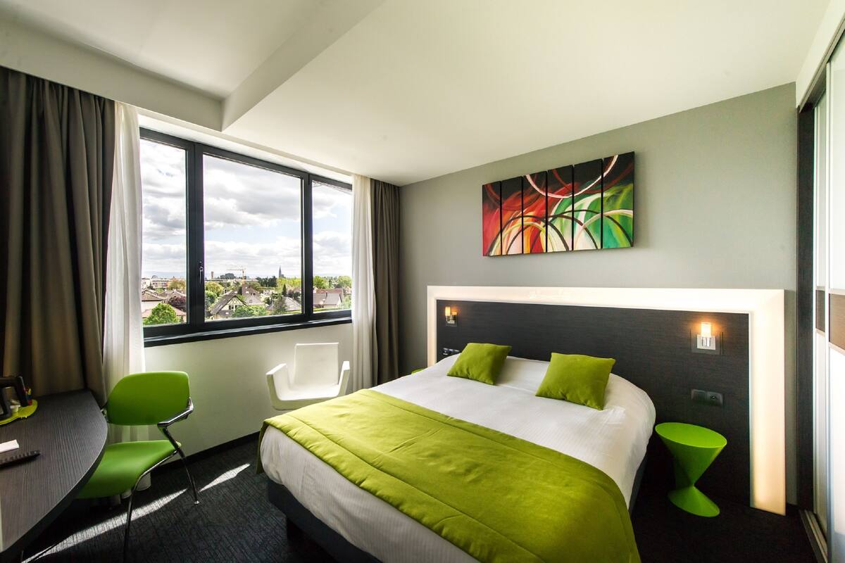 A hotel room with a large bed, green pillows, a desk with a chair, a small green side table, and a painting on the wall.