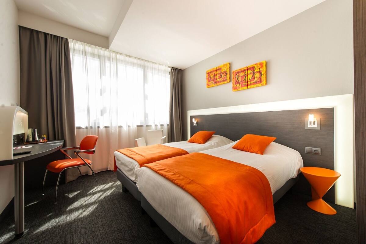 A hotel room with two beds, a desk, a chair, and a large window with curtains.
